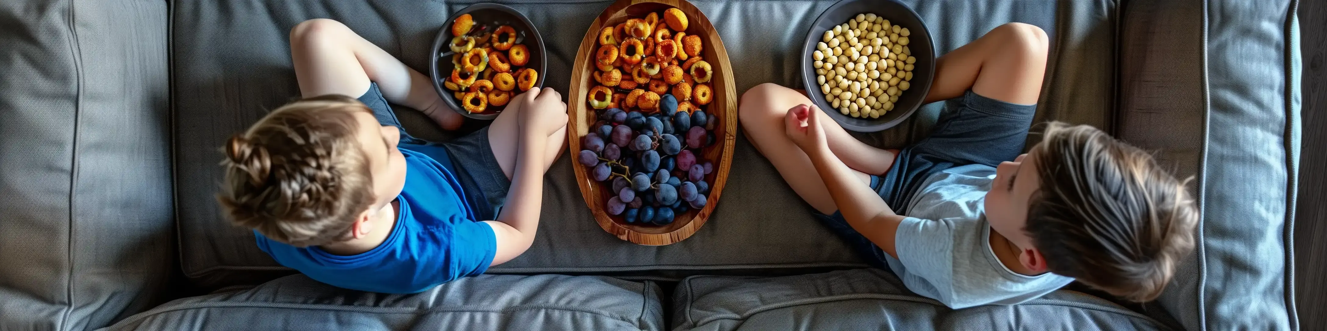 Zwei Kinder sitzen auf einem Sofa, zwischen ihnen Schalen mit Snacks und Trauben, von oben gesehen