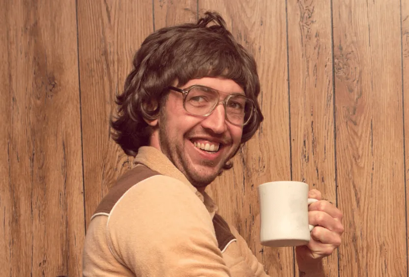Un hombre con melena setentera y gafas grandes sonríe con una taza de café